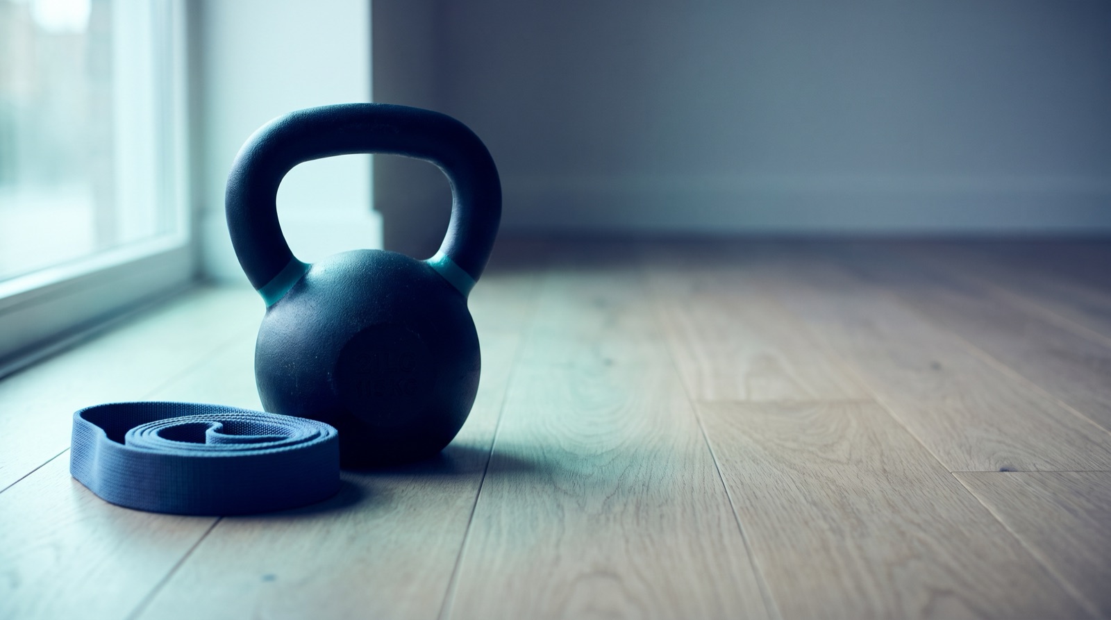 A dark kettlebell and a coiled blue resistance band on a pale wood floor beside a window.