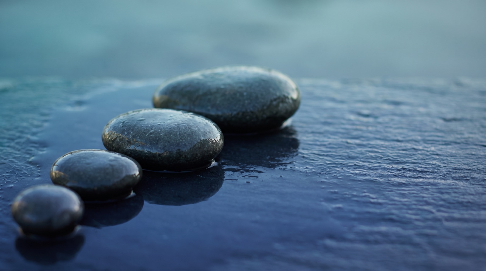 Four smooth dark stones, each larger than the last, lined up on a wet slate surface under cool blue light.