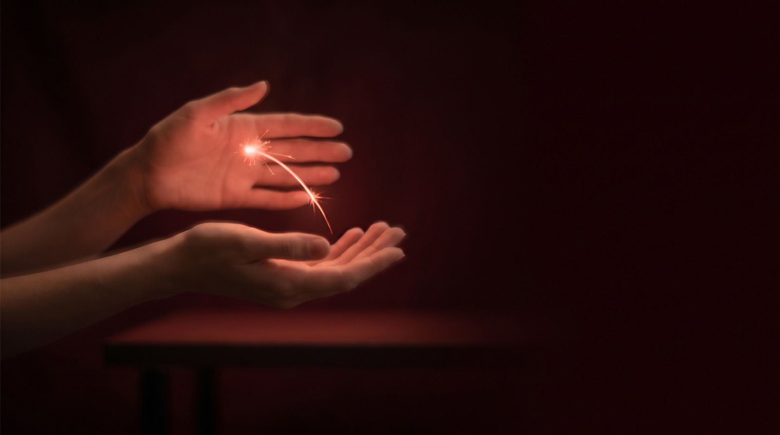 Two open hands, one held above the other in warm red light, with a small spark of light arcing between the palms