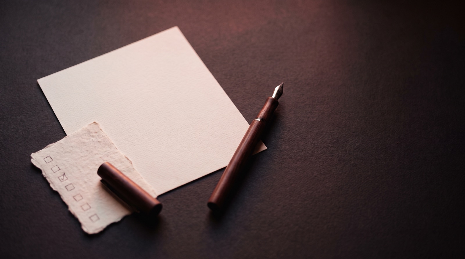 A blank sheet of paper, a small torn checklist with one box ticked, and a fountain pen resting on a dark desk