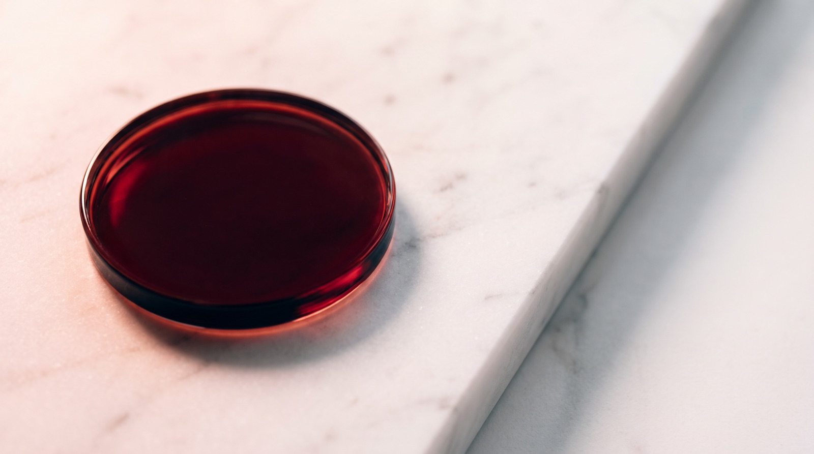 A shallow round glass dish holding dark red liquid, sitting on a white marble slab.
