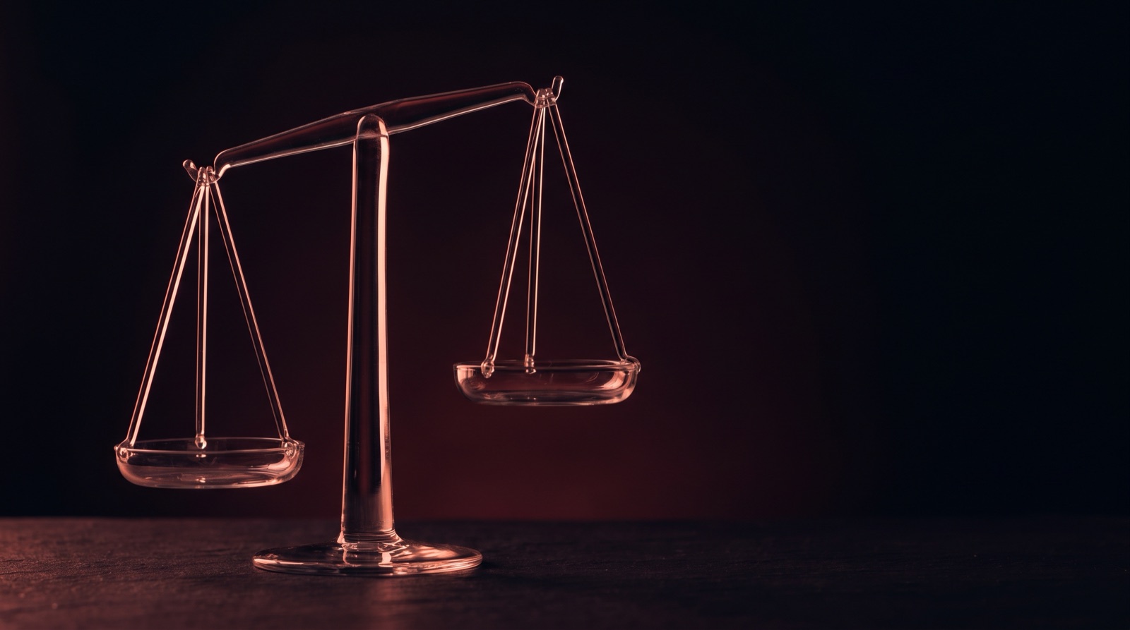 A small glass balance scale tipped out of level, one pan hanging lower than the other, against a dark red background.