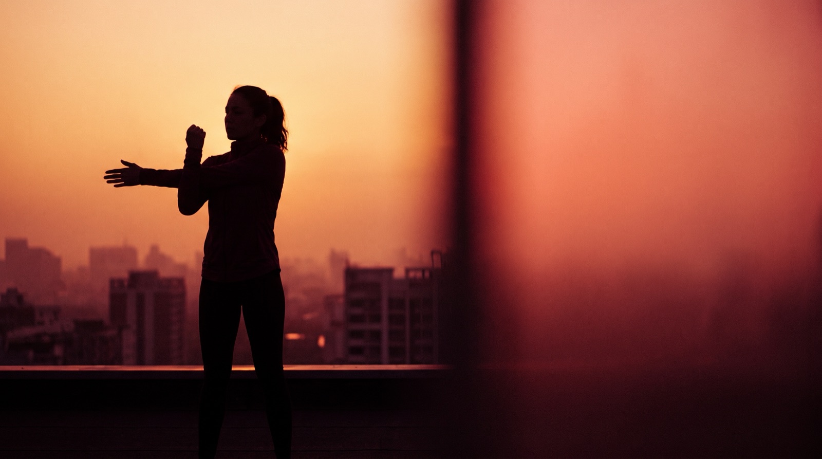 The silhouette of a person stretching one arm across the chest on a rooftop at sunrise, with a city skyline behind them