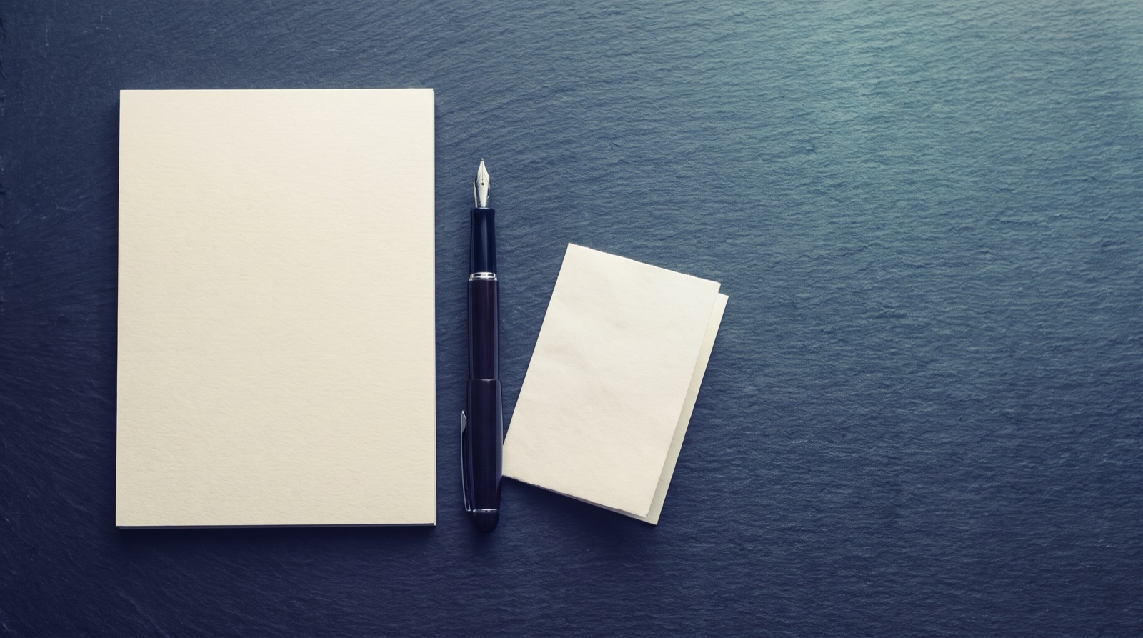 A blank notepad, a fountain pen, and a small folded blank card laid flat on a dark slate surface.